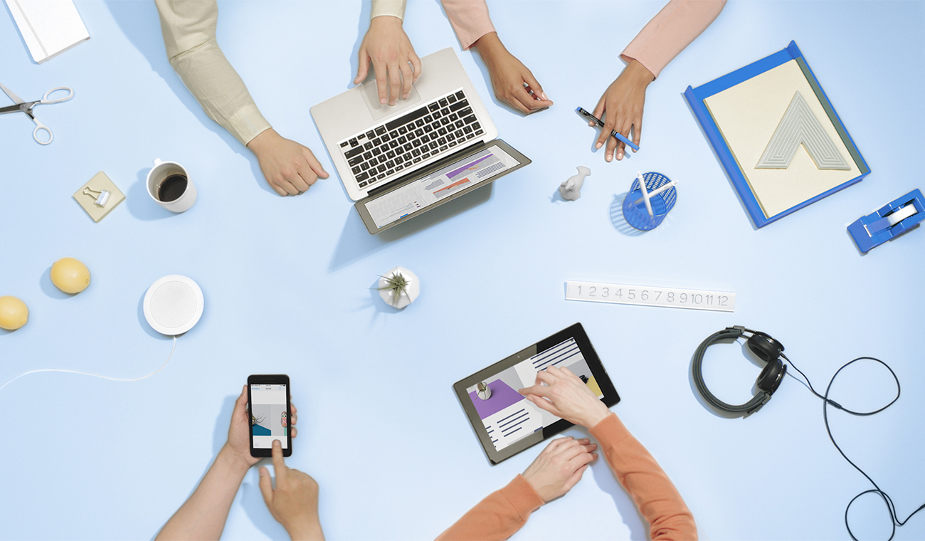 Overhead view of several people working on computers, tablets, and phones with their hands.