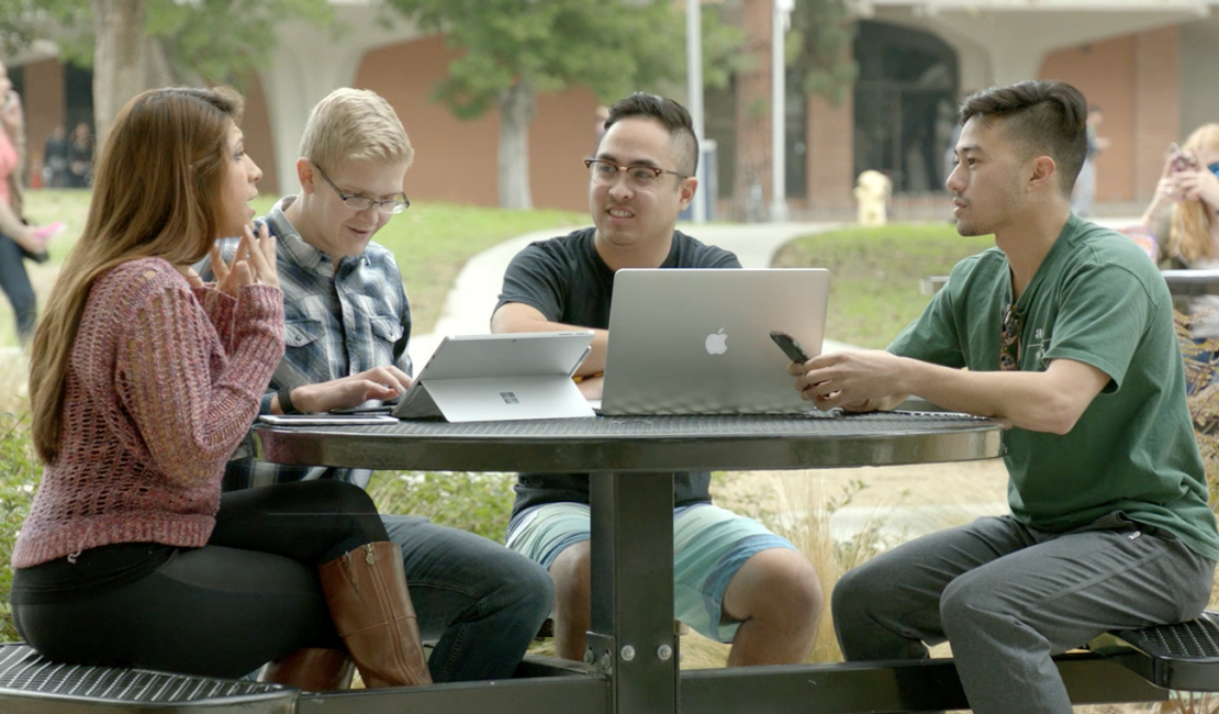 Four college students sitting around a picnic table outside, talking over laptops and tablets.