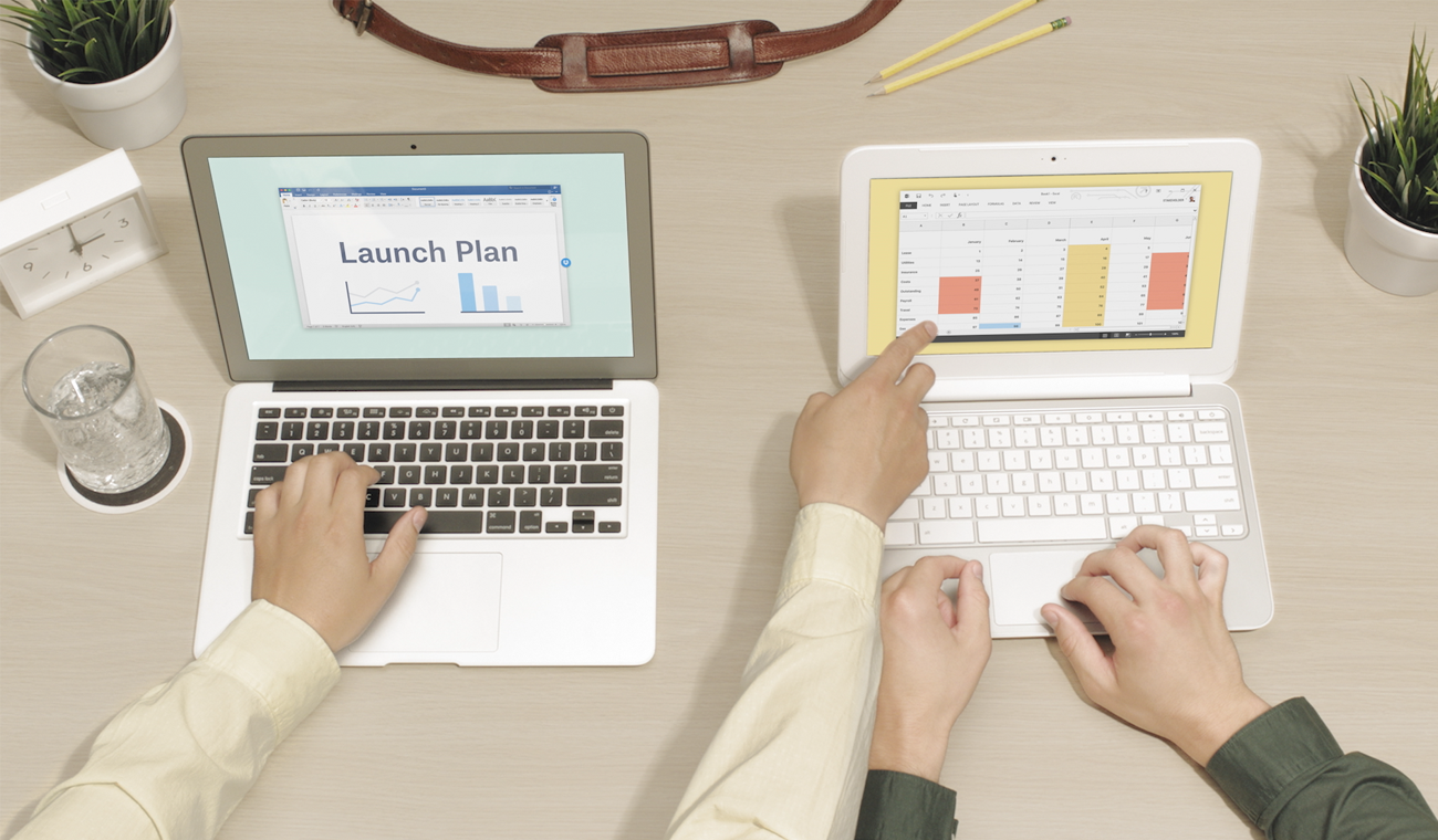 Two people working side-by-side on two laptops. The person on the left is pointing out something on the laptop screen of the person on the right.