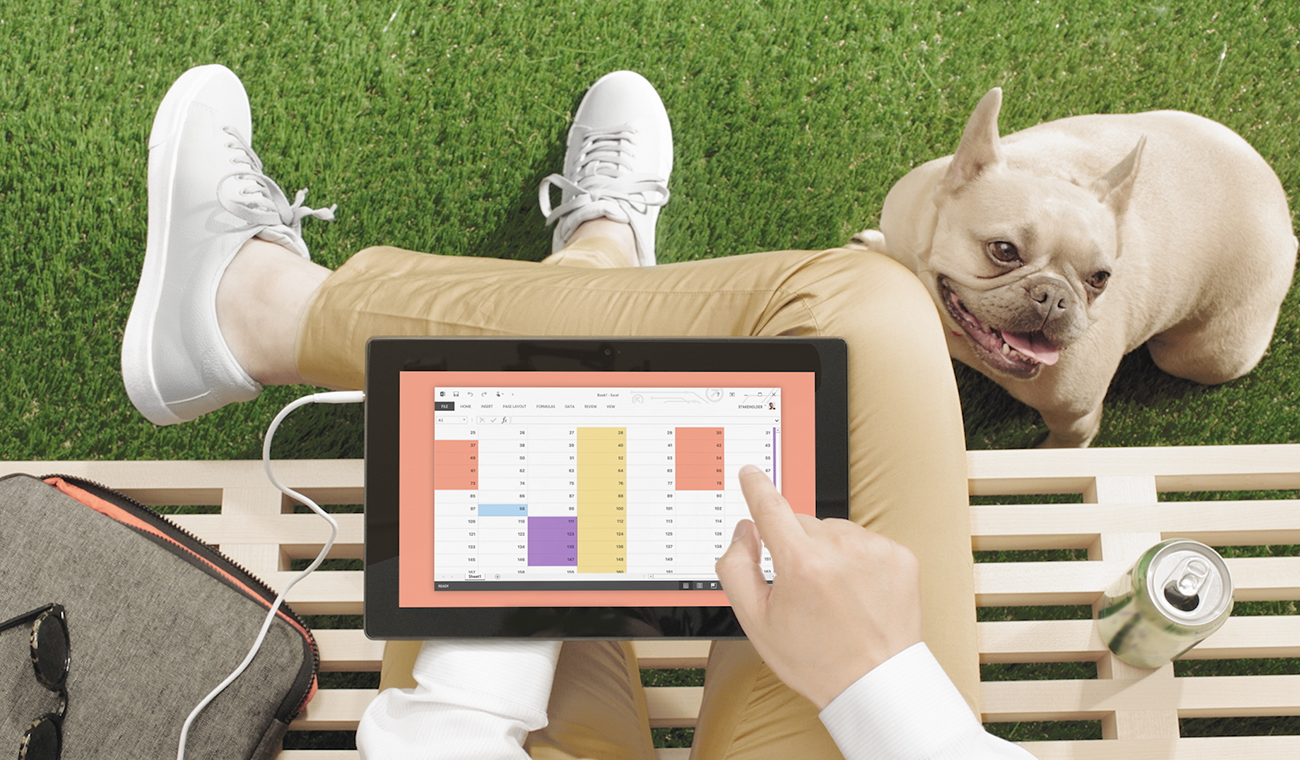 A person sitting on a park bench, using a calendar app on a tablet, with a dog sitting nearby on the grass.