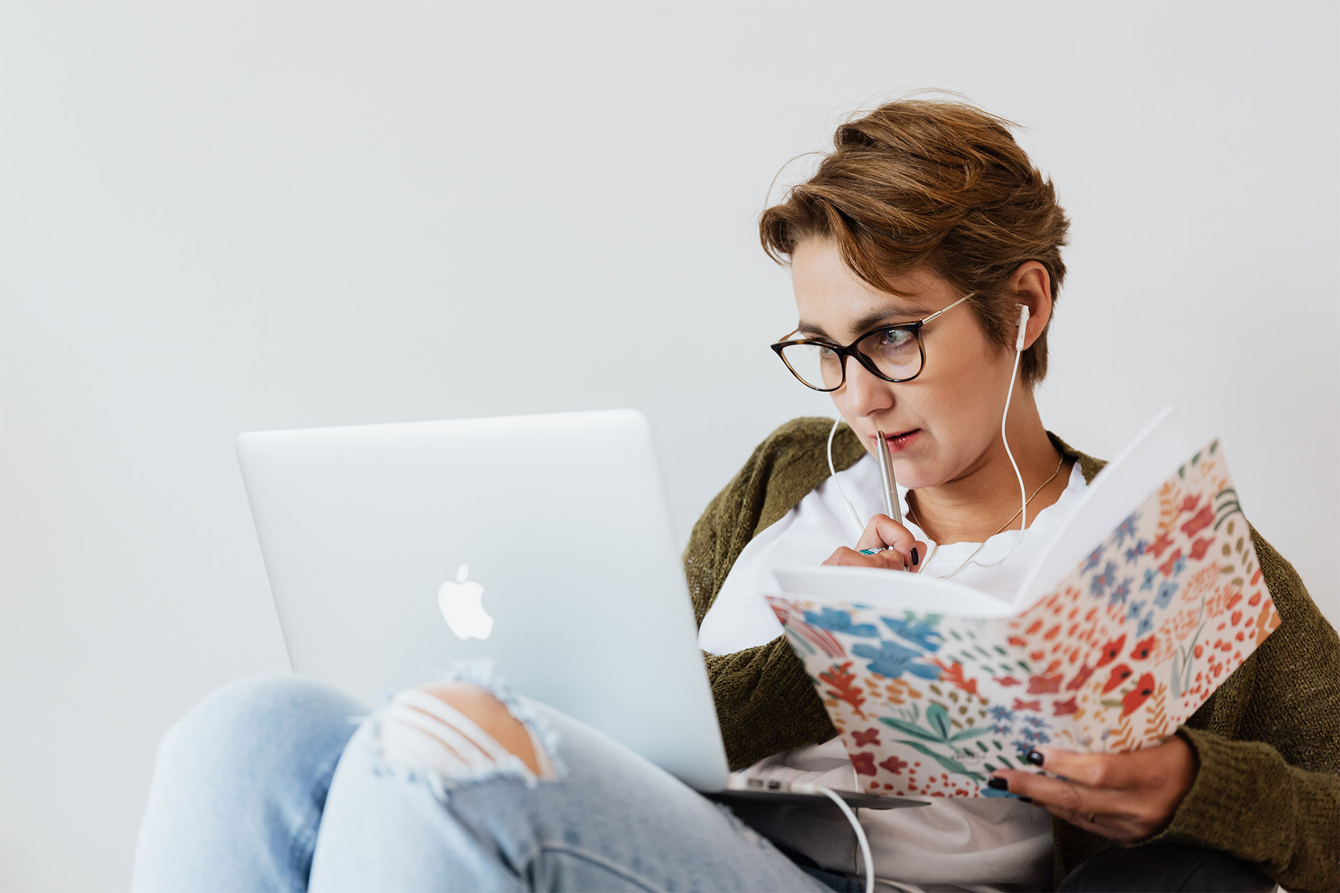 イヤフォンをして MacBook で作業をしている人物。手にはノートとペンを持っている