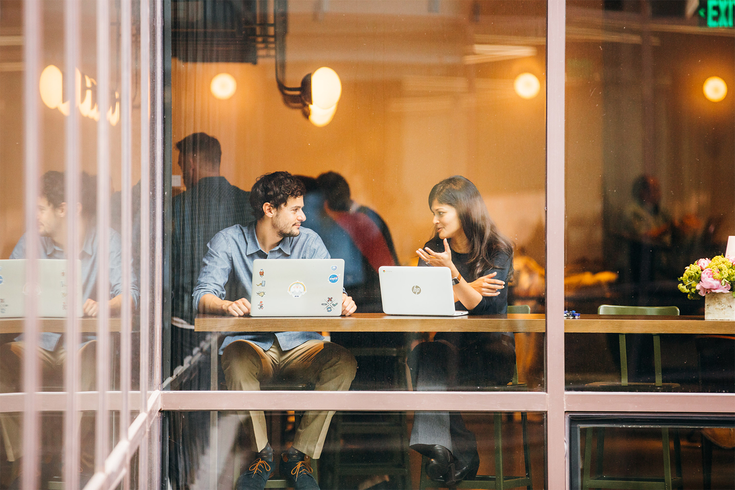 Two people working on laptops have a conversation.