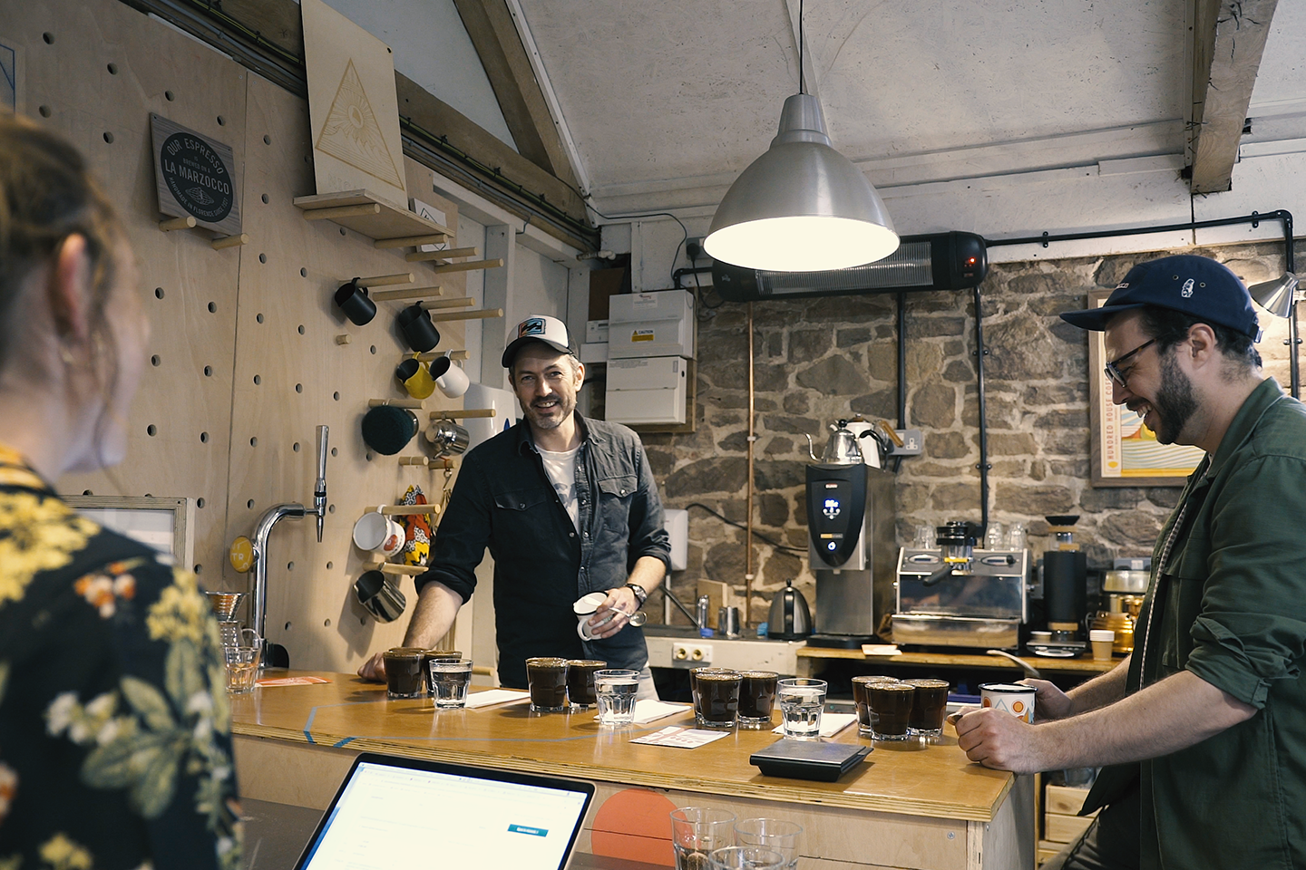 Two men sample coffee while a woman works on a laptop.