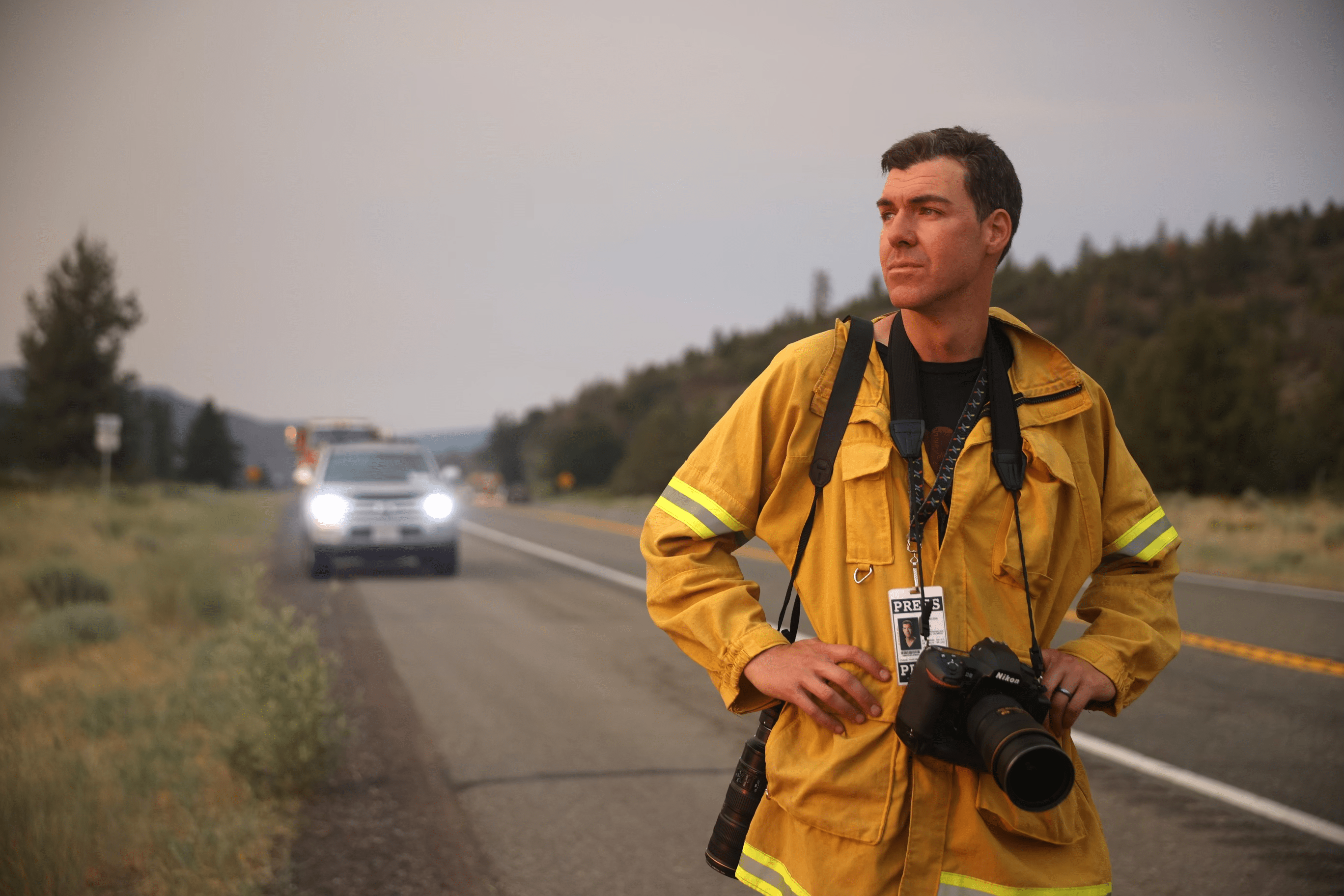 Photographer Josh Edelson surveys the landscape while standing in the road with a camera and press badge around his neck.