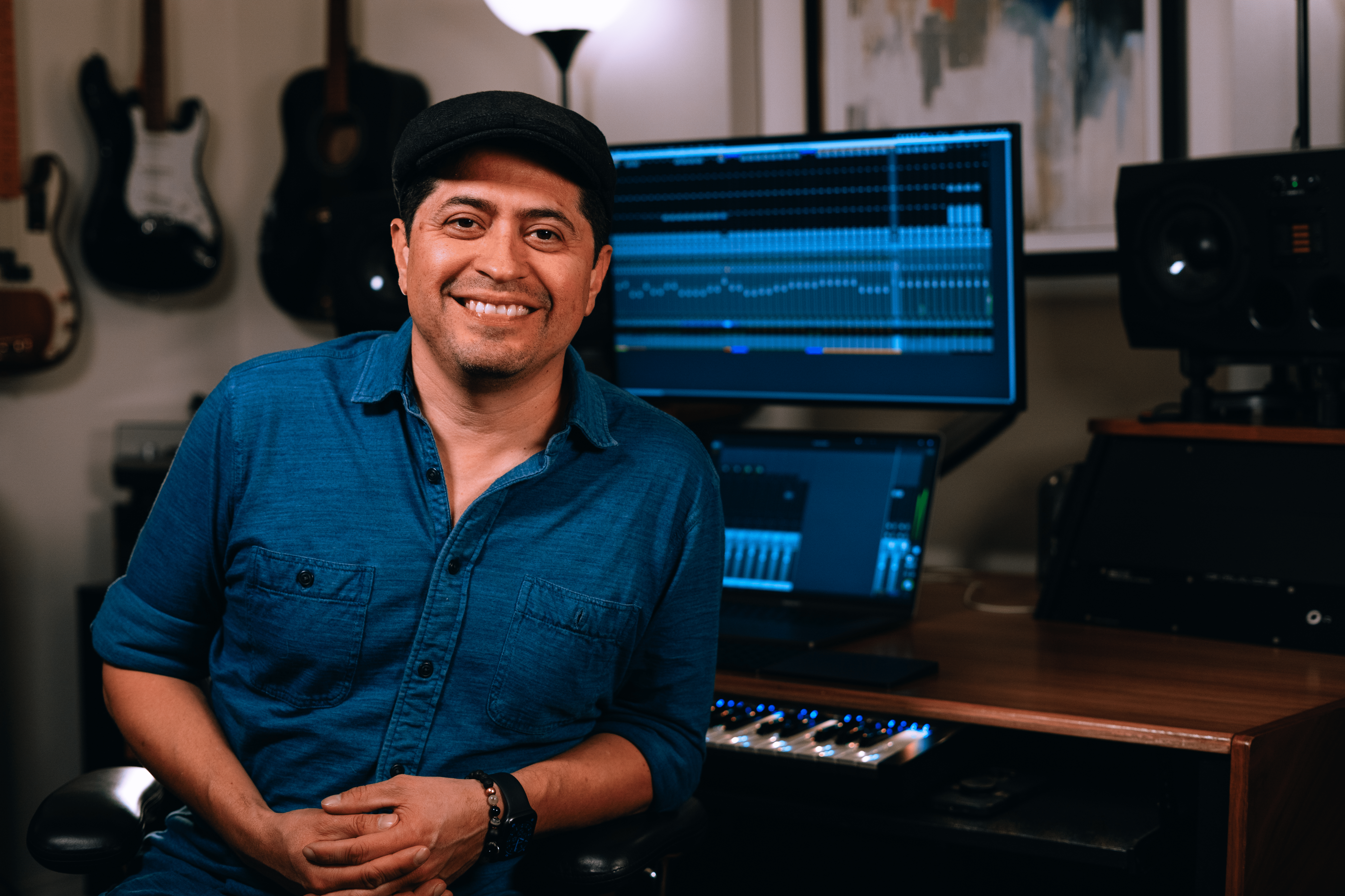 O compositor, produtor e engenheiro Miguel Soltero está sentado e sorrindo em seu estúdio com guitarras ao fundo.