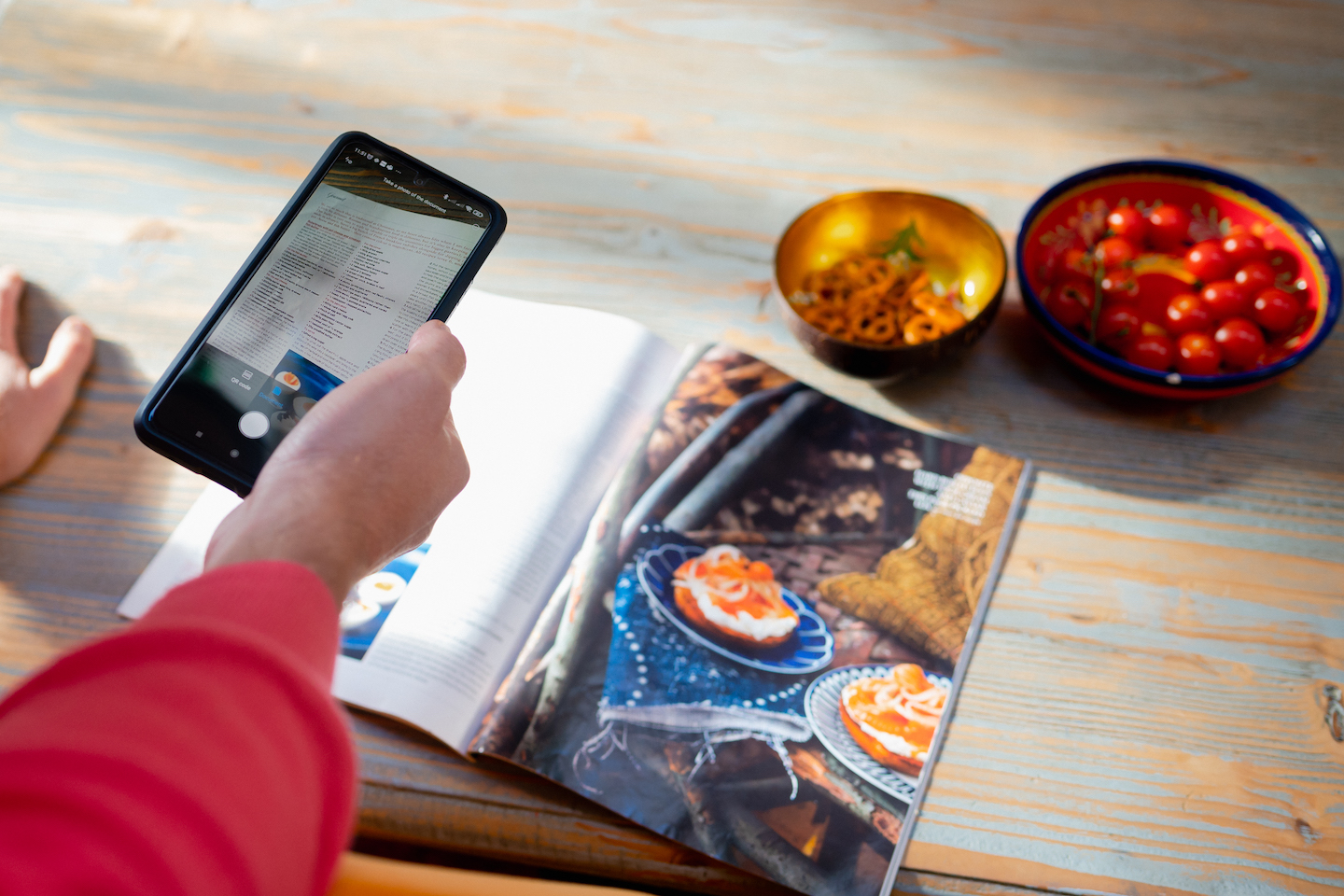 使用手机拍摄杂志上的食谱，旁边放有装着西红柿和椒盐脆饼的小碗