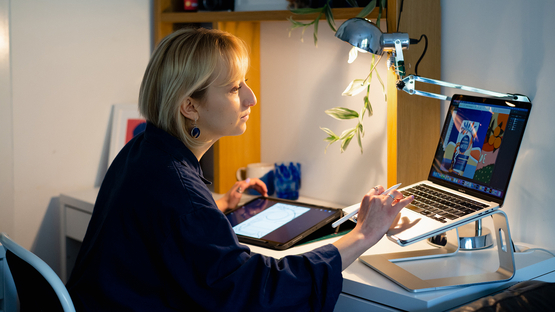 Femme assise à un bureau, tapant sur un ordinateur et tenant un stylet à la main, et tablette visible au second plan