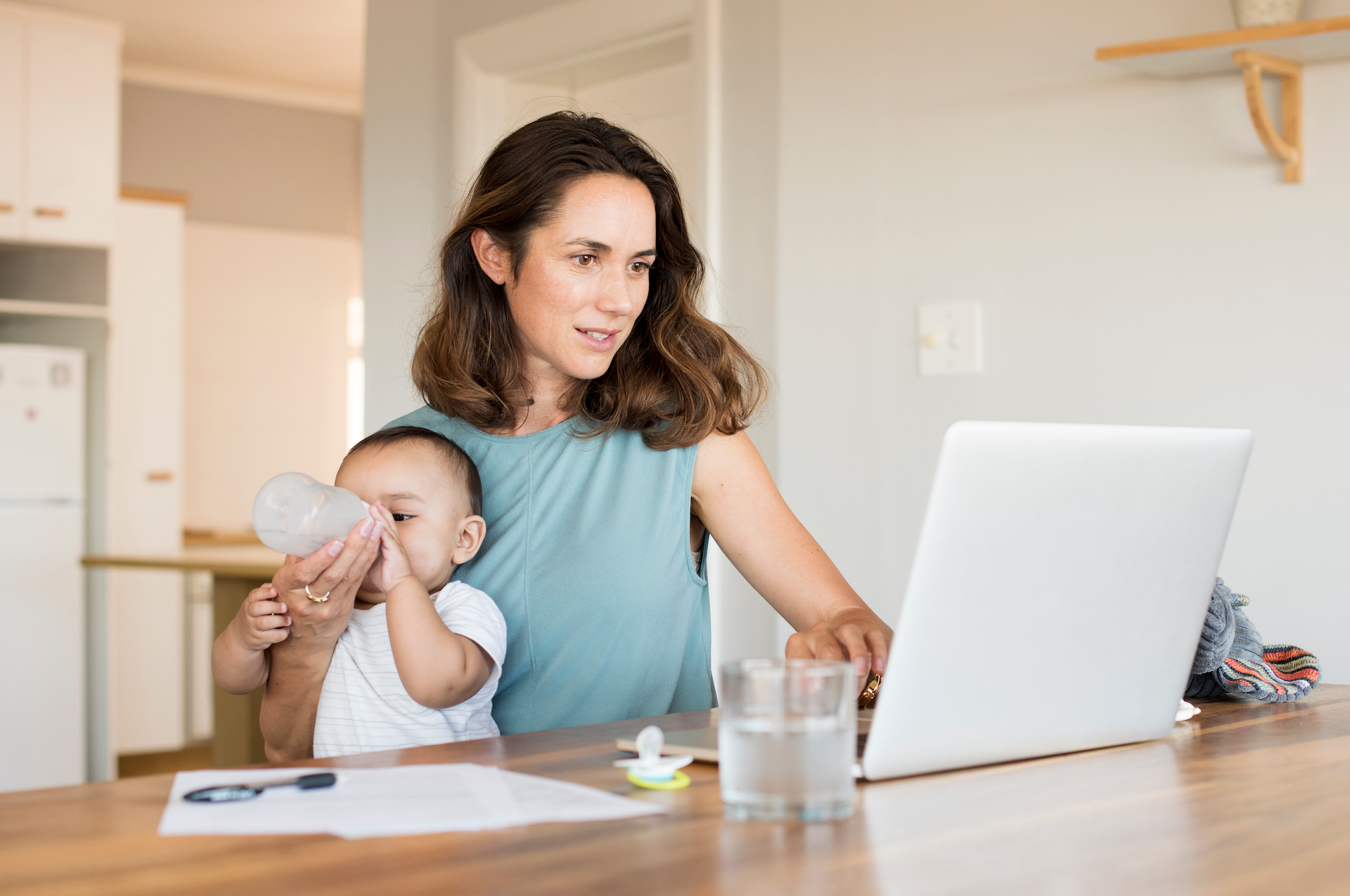 Woman works on a laptop while holding and feeding a baby in her kitchen