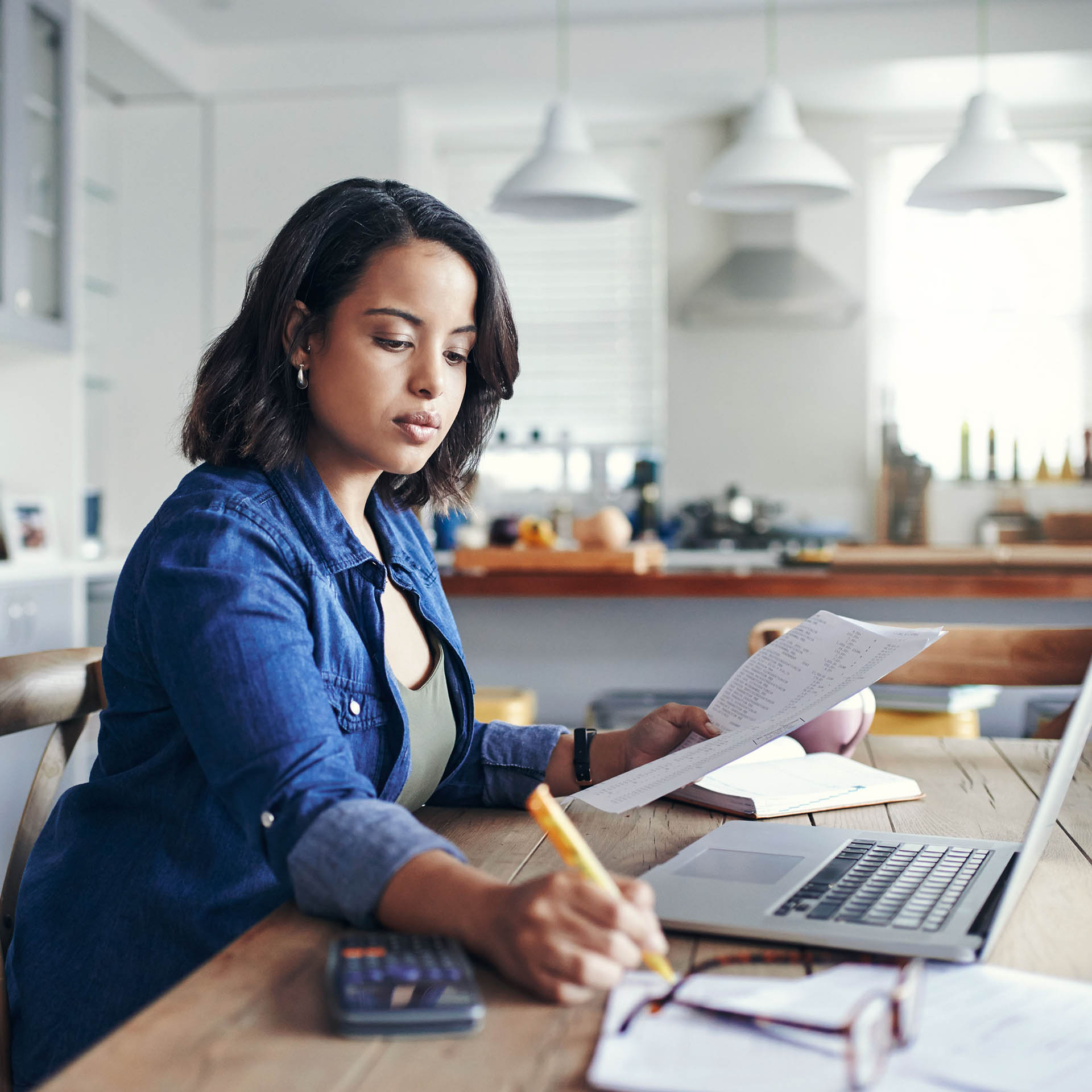 En kvinne skriver på papir mens hun sitter ved en kontorpult foran en bærbar datamaskin