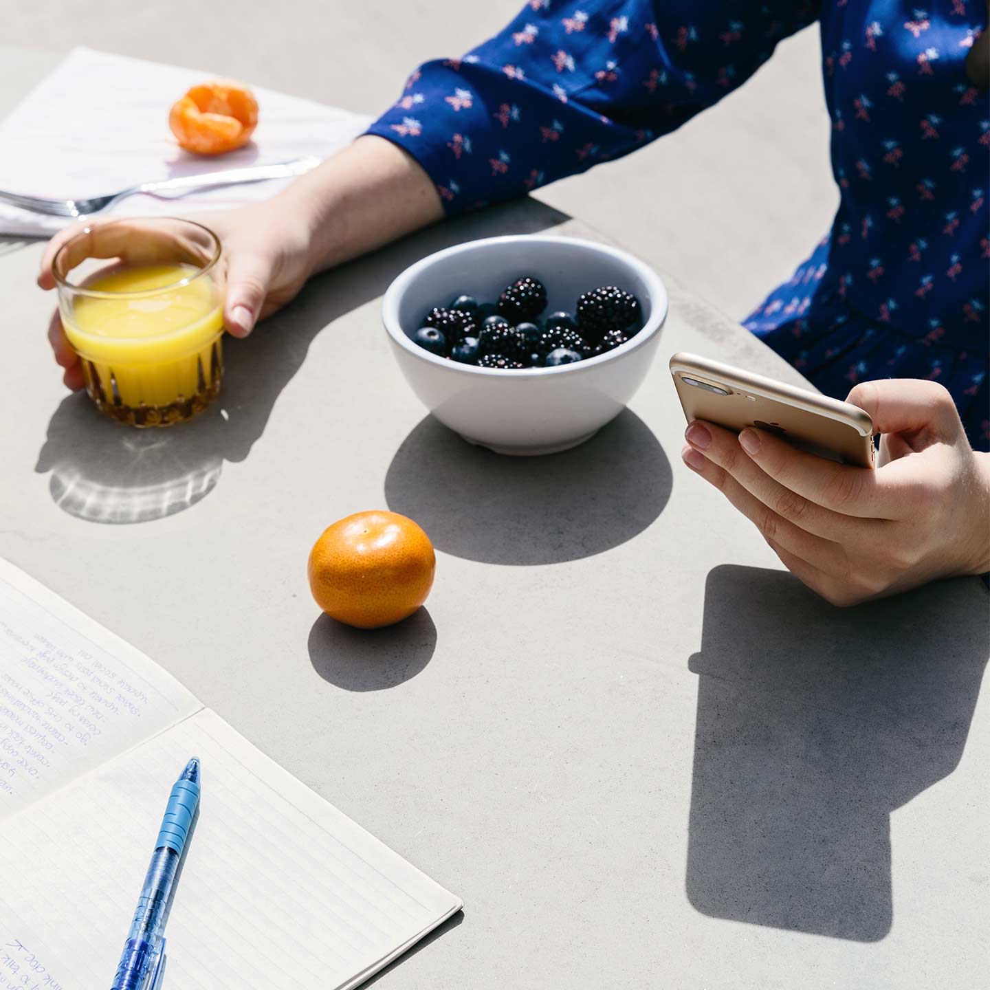 Seseorang sedang memegang ponsel dan segelas jus jeruk di depan mangkuk berisi buah beri