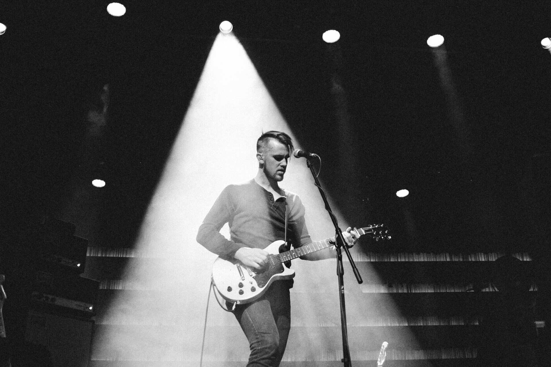 音乐人 Peter Ferguson 站在舞台上的聚光灯下，背着吉他面对话筒