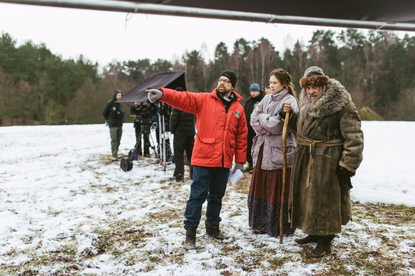 Outdoor winter film shoot with camera equipment; in foreground 1 crew member points next to 2 actors in large coats 