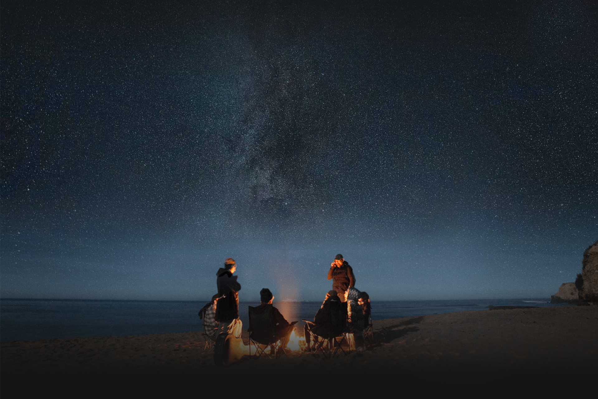 Sieben Personen sitzen und stehen in einer sternenklaren Nacht um ein Lagerfeuer am Strand