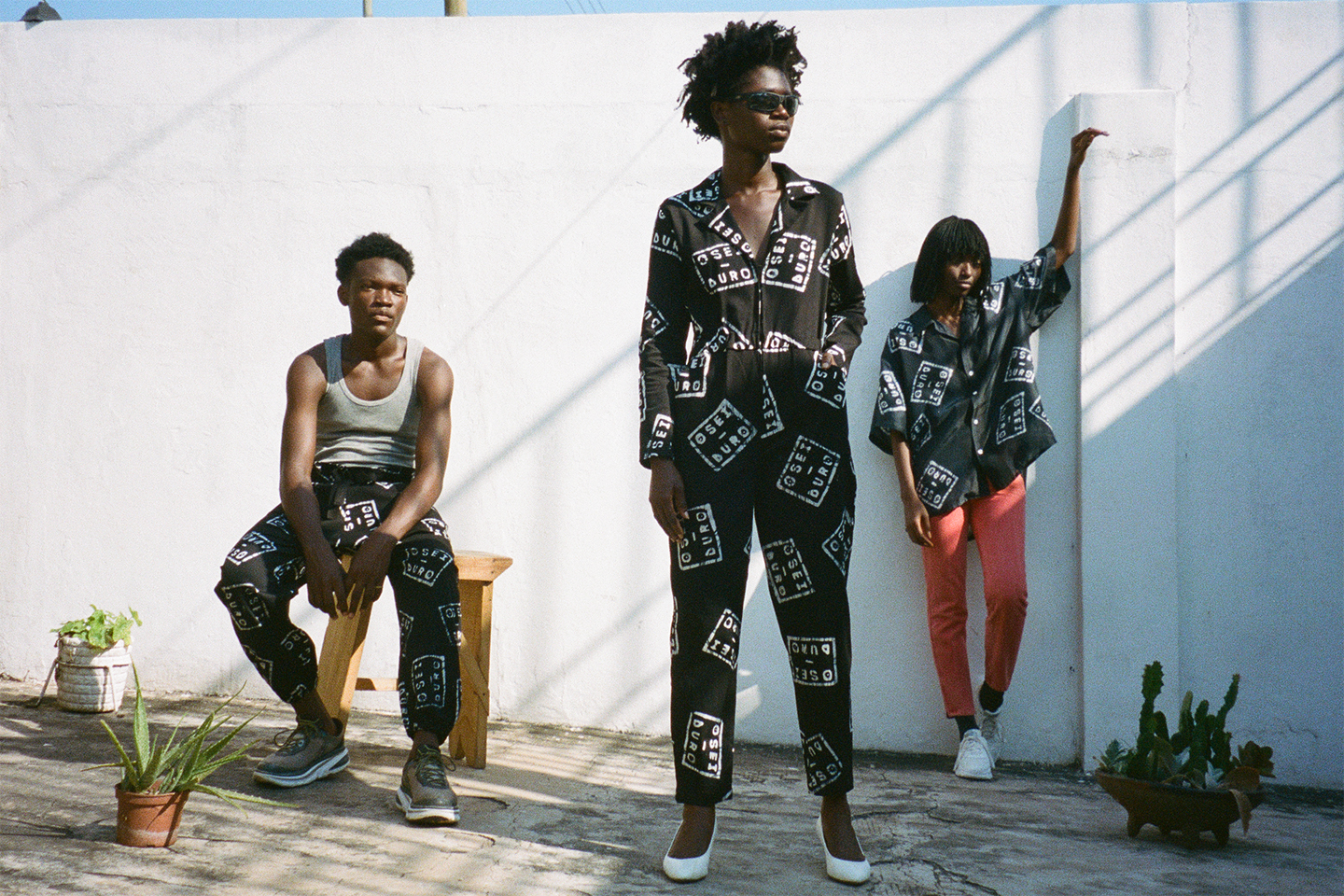 3 people in a courtyard with 3 potted plants wear pants, jumpsuit, shirt with OSEI DURO print
