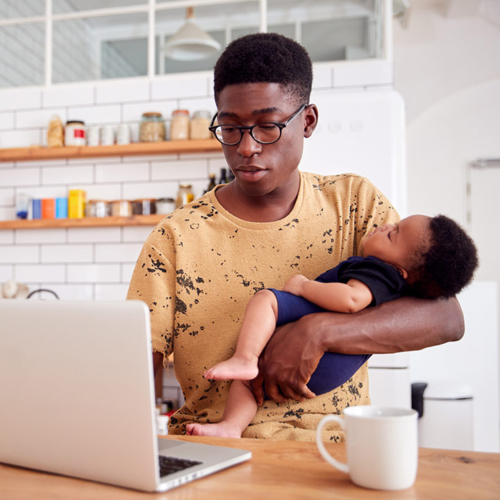 A parent works from home on a laptop while holding their child