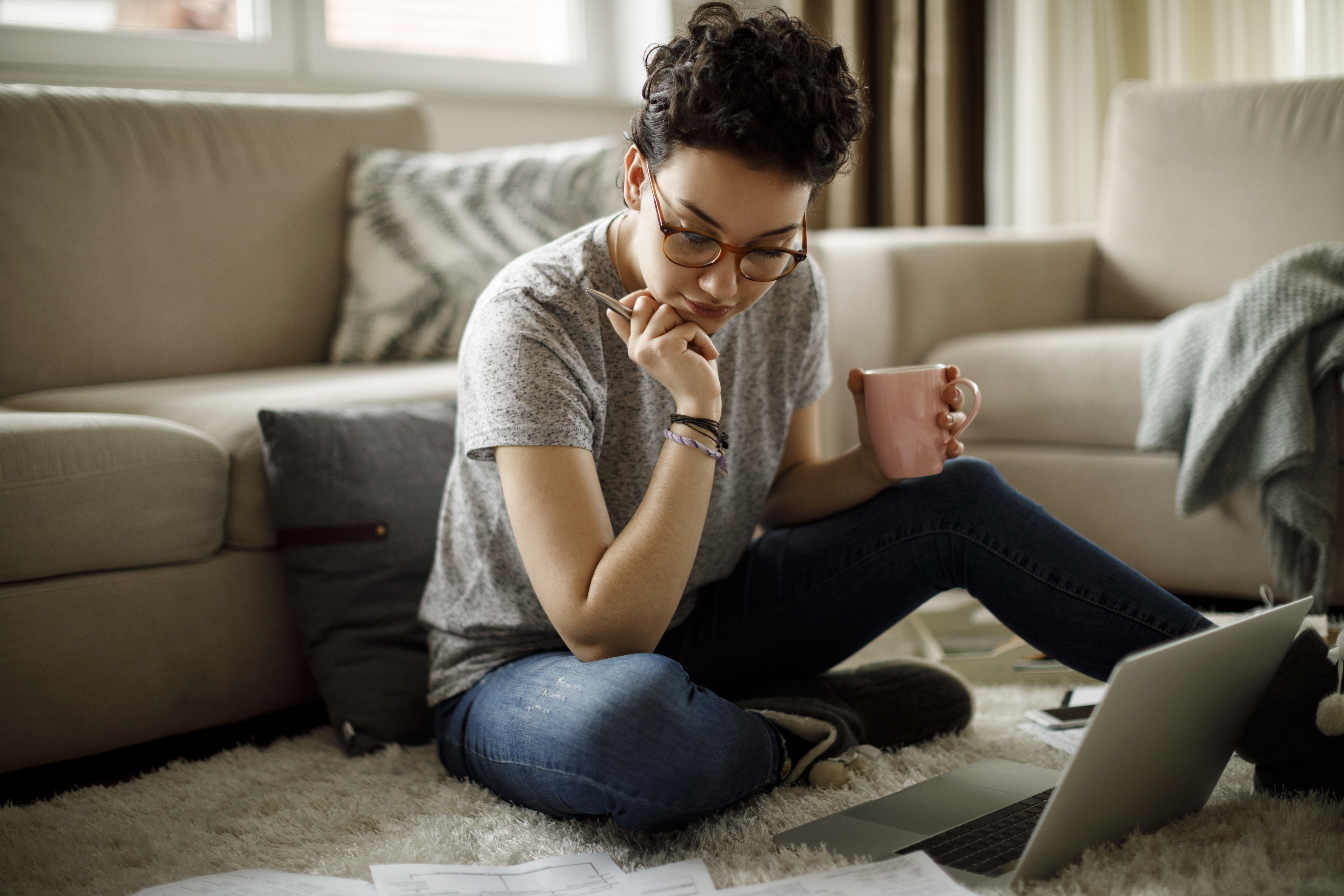 A freelancer working at home sits on the floor and looks at some papers while their laptop is open in front of them.