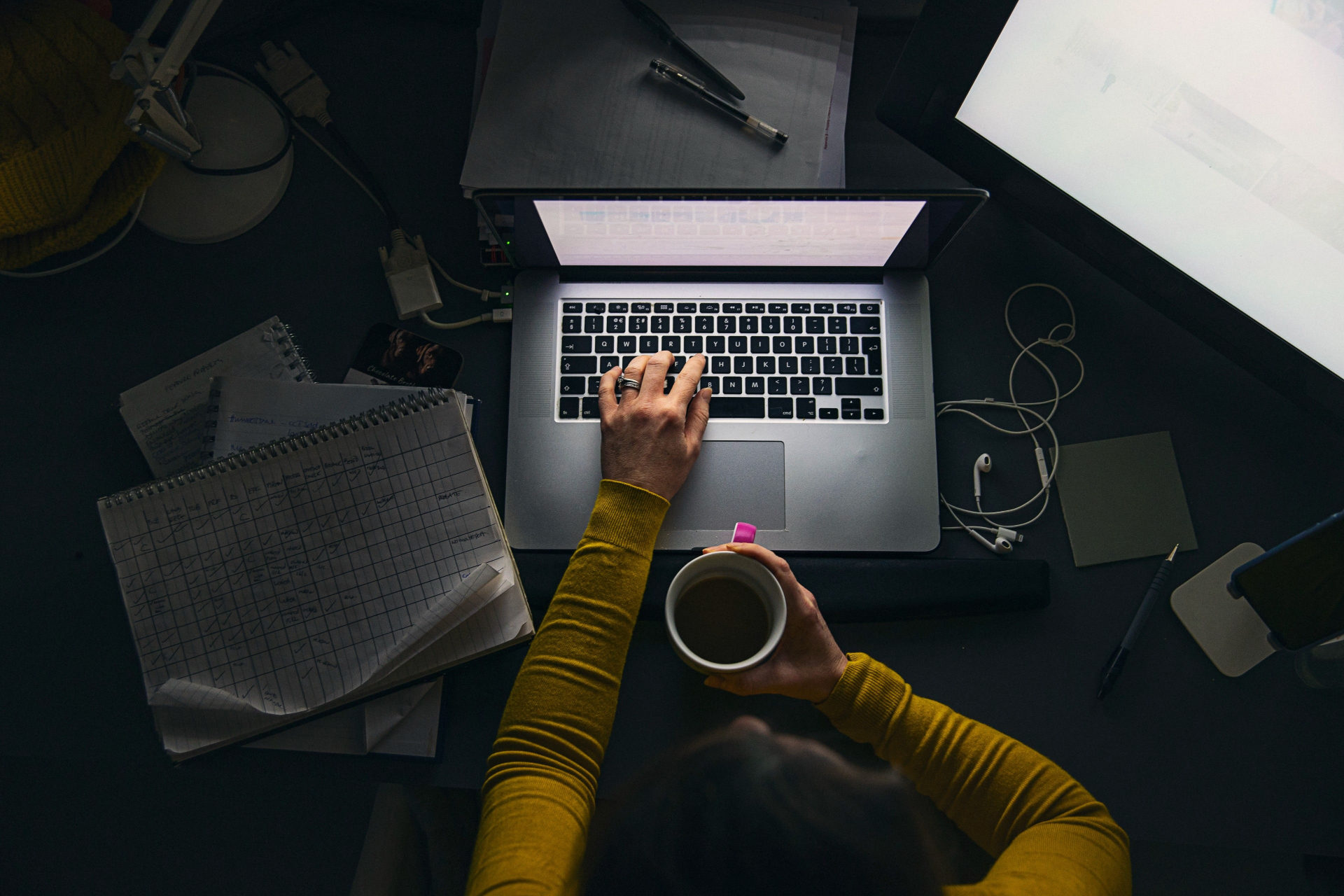 Persoon die aan een bureau zit, op de laptop werkt en een kopje koffie vasthoudt