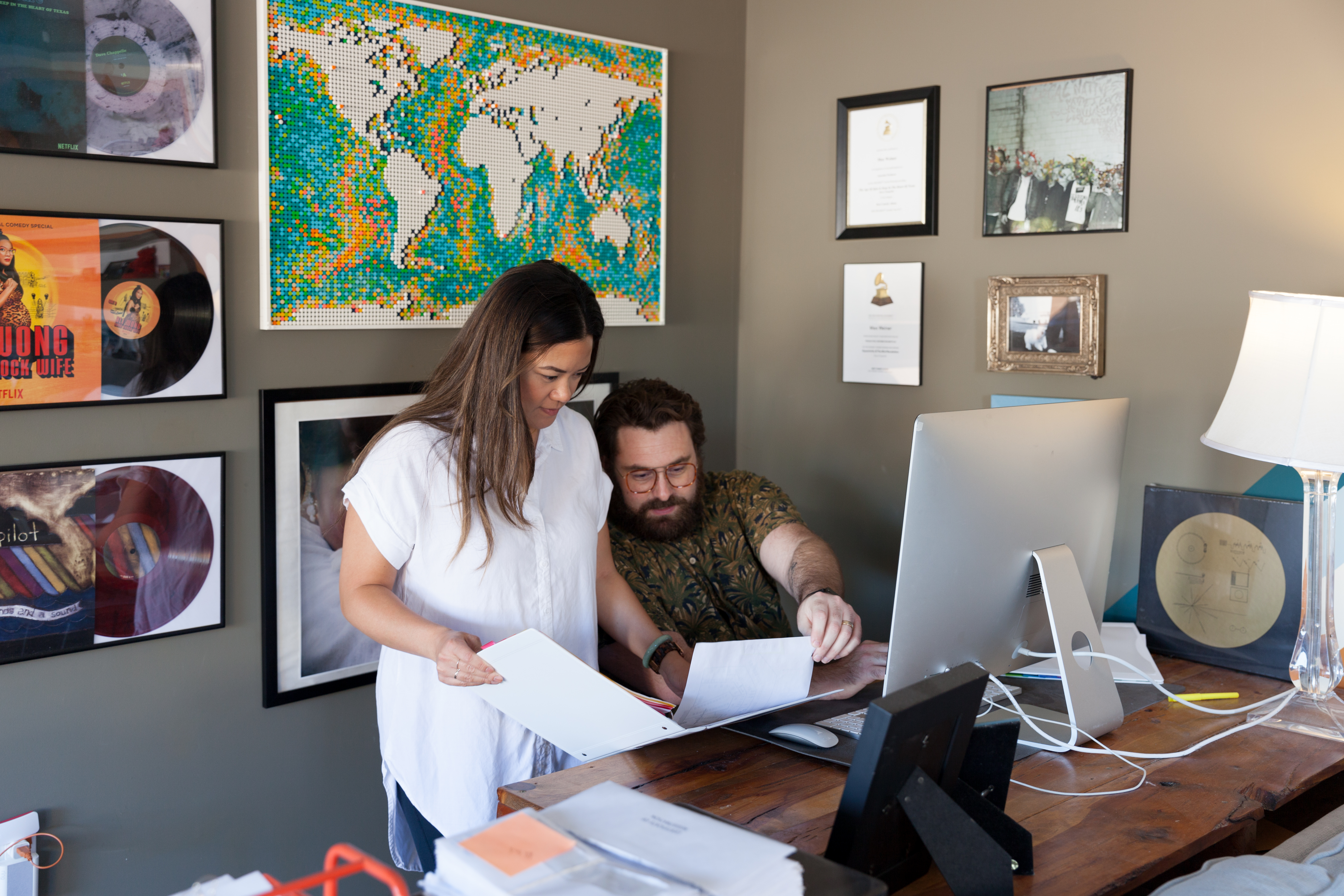 Two creative professionals work at a monitor while reviewing project documents.