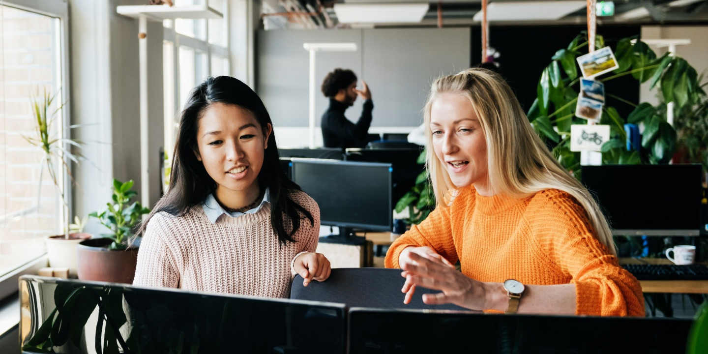 Two women in a modern office, seated at a desk in front of a computer monitor.