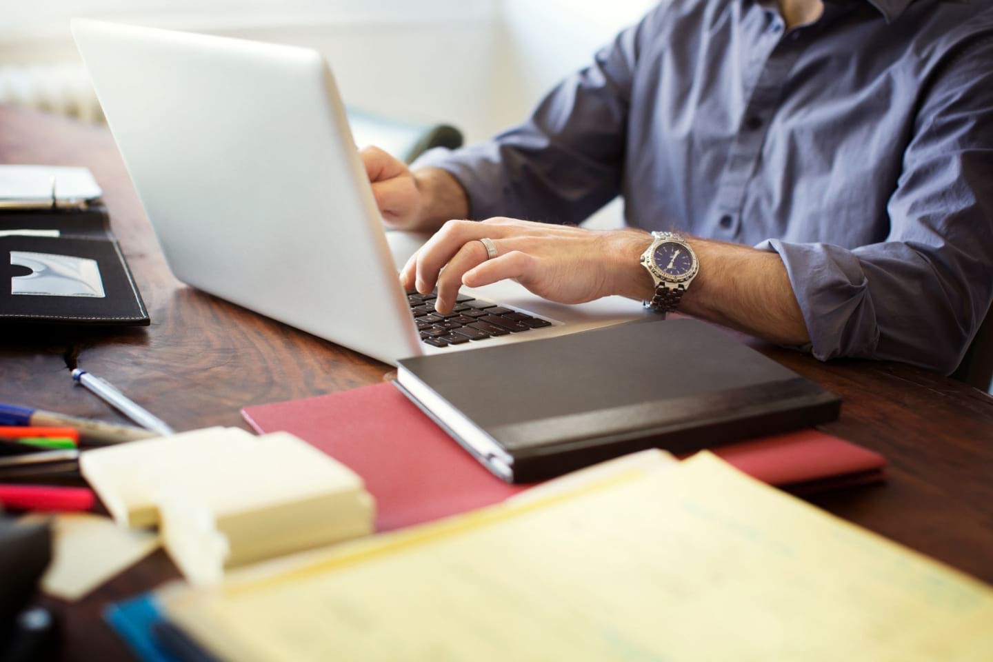 Osoba pracuje na laptopie przy biurku, na którym leżą długopisy, karteczki samoprzylepne i notatniki.