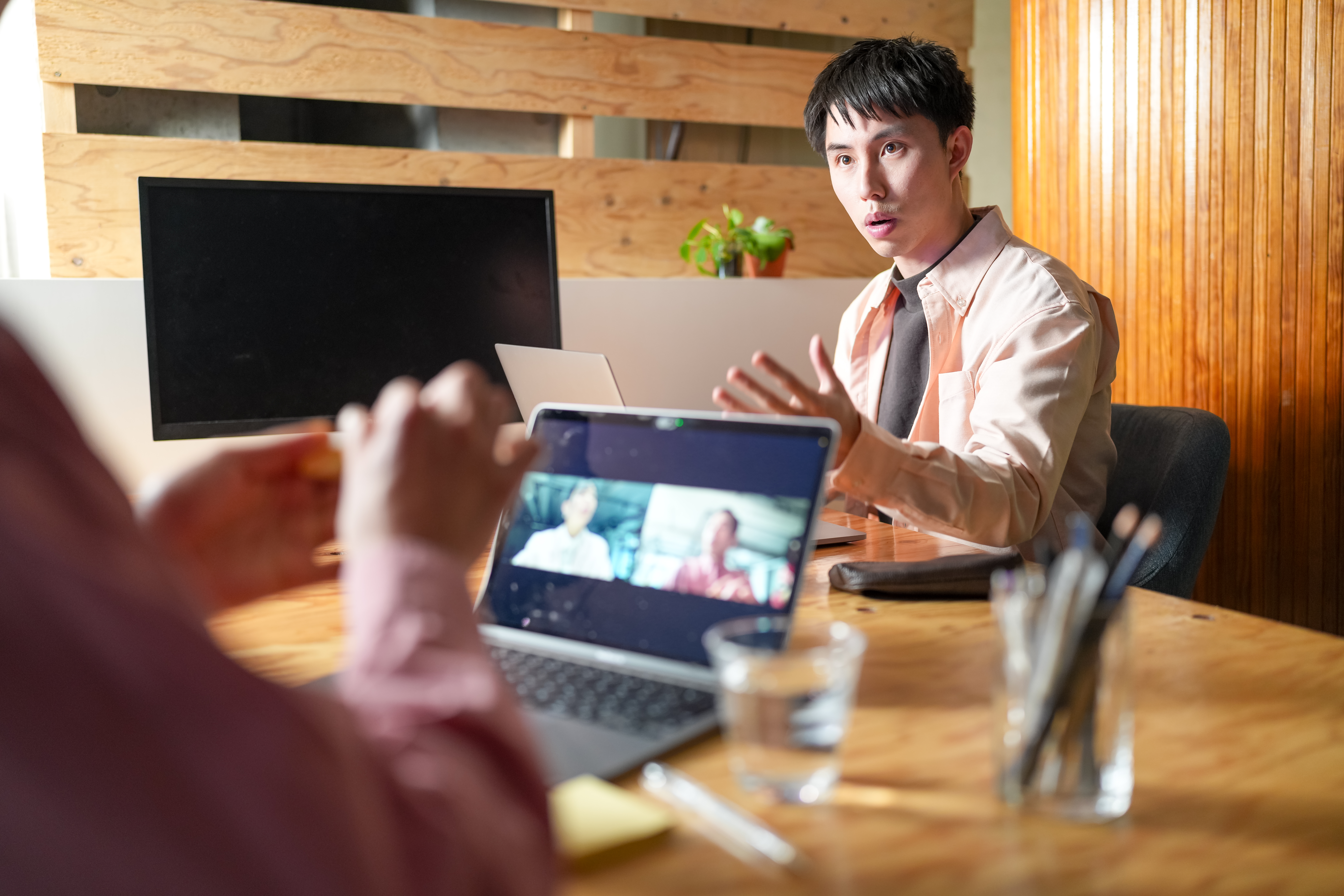 Person in a meeting speaking to a colleague who is seen through a laptop screen in a video call setup.