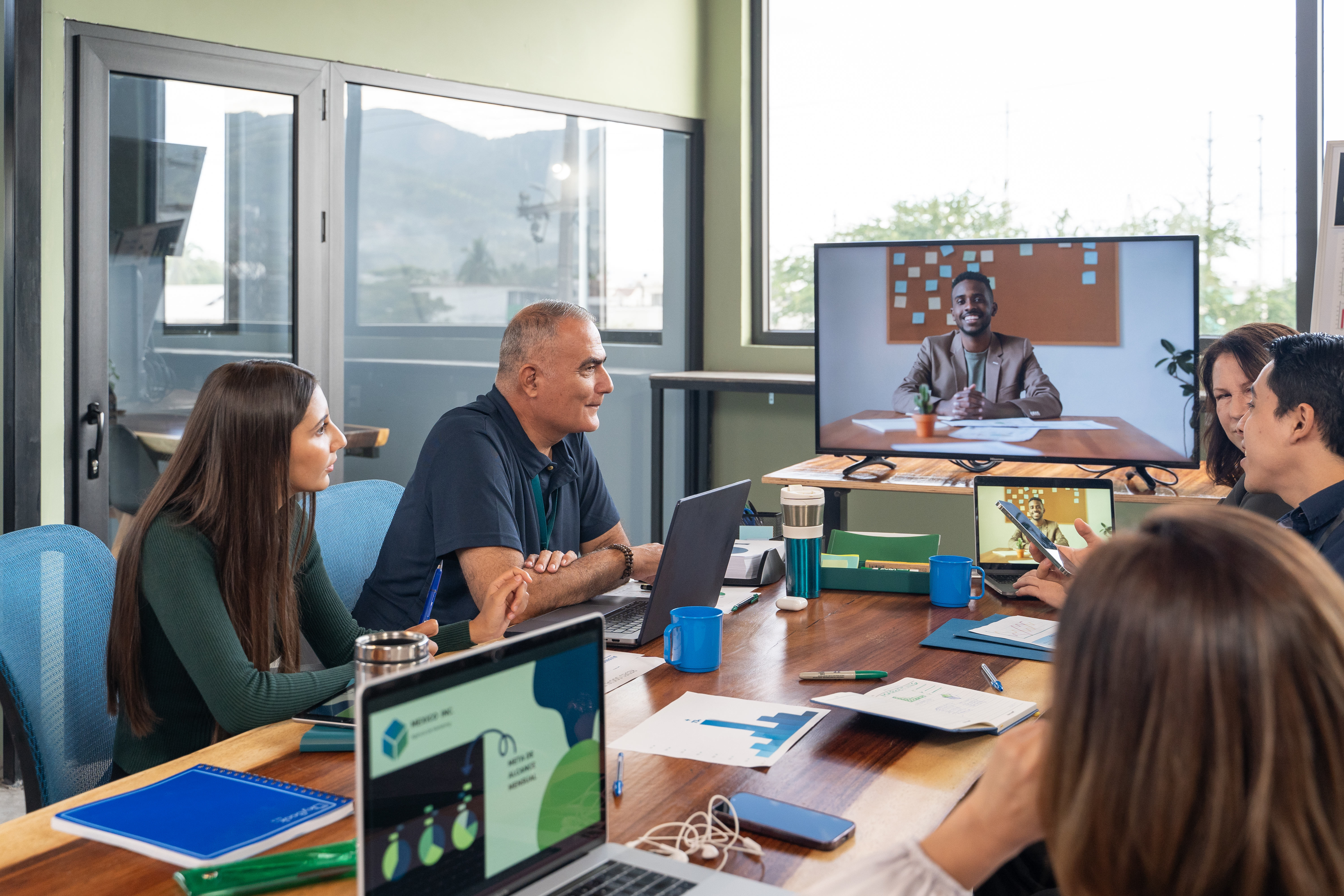 Group of people in an office participates in a video call with a remote colleague displayed on a large screen.