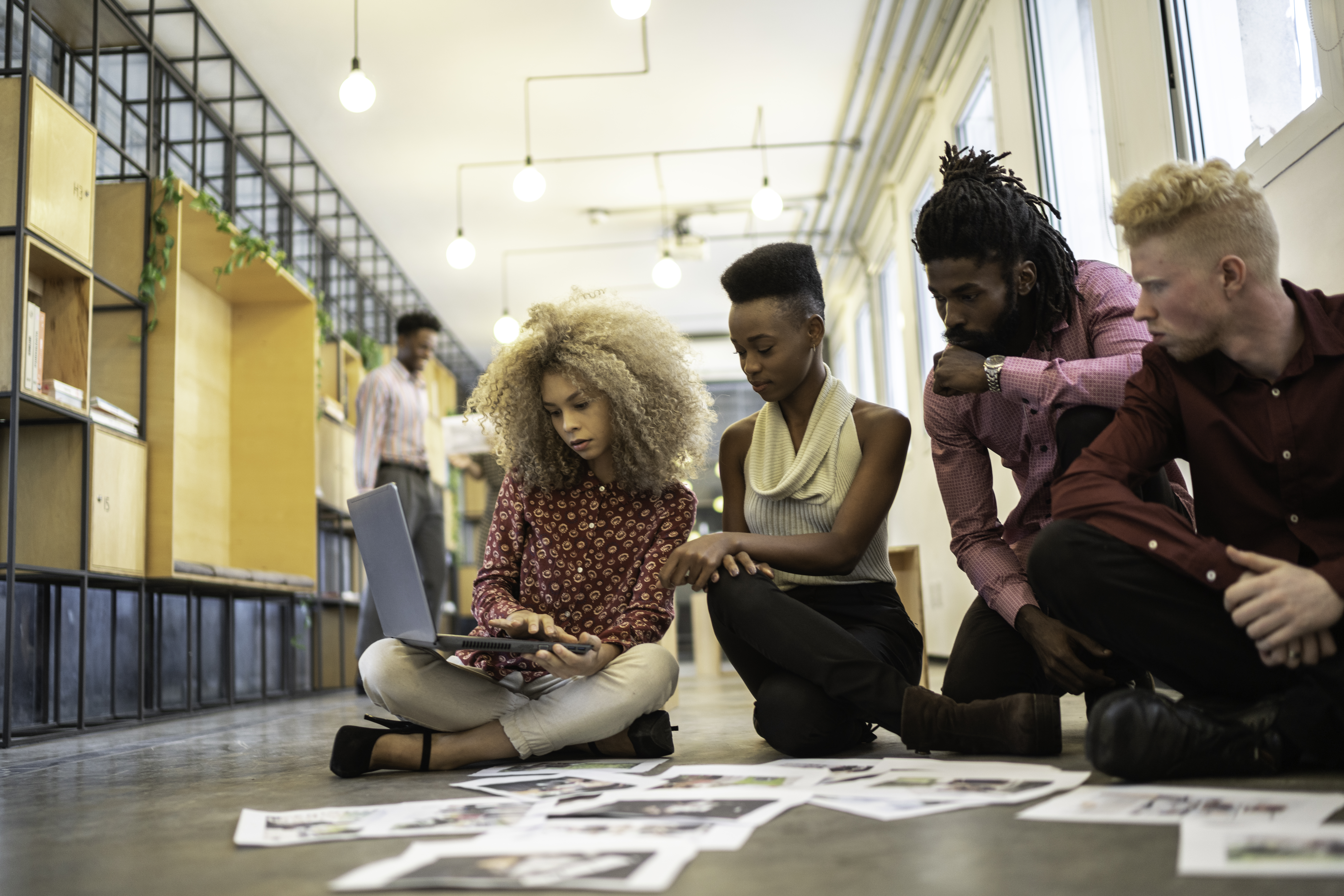 Group of people sitting on the floor reviewing images on a laptop and scattered papers in a modern office space.