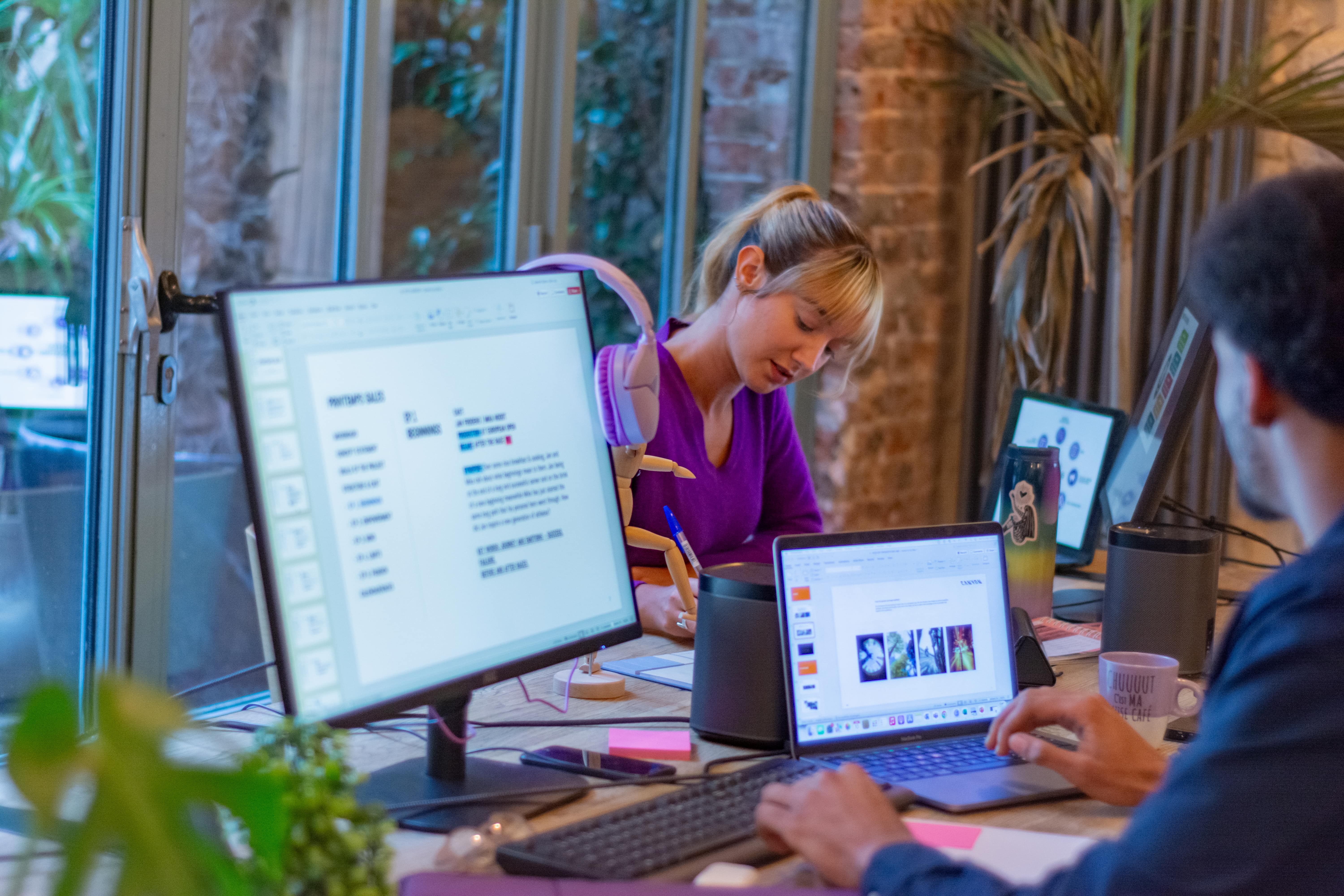 Two people work at a desk with laptops and monitors, one person is working on slides, and the other is writing.