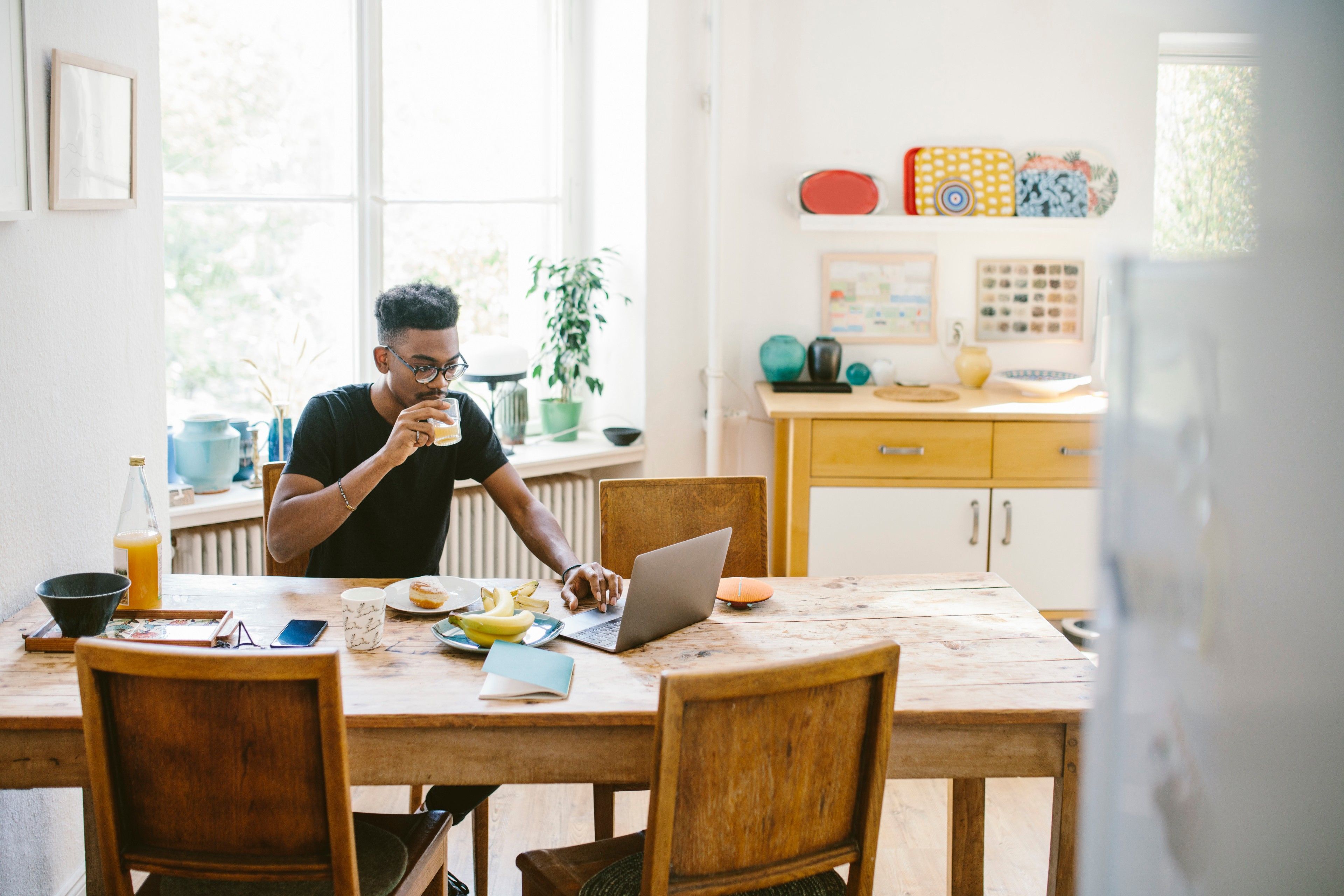 Personne travaillant depuis chez elle, assise à une table de cuisine, et buvant une boisson en consultant son ordinateur