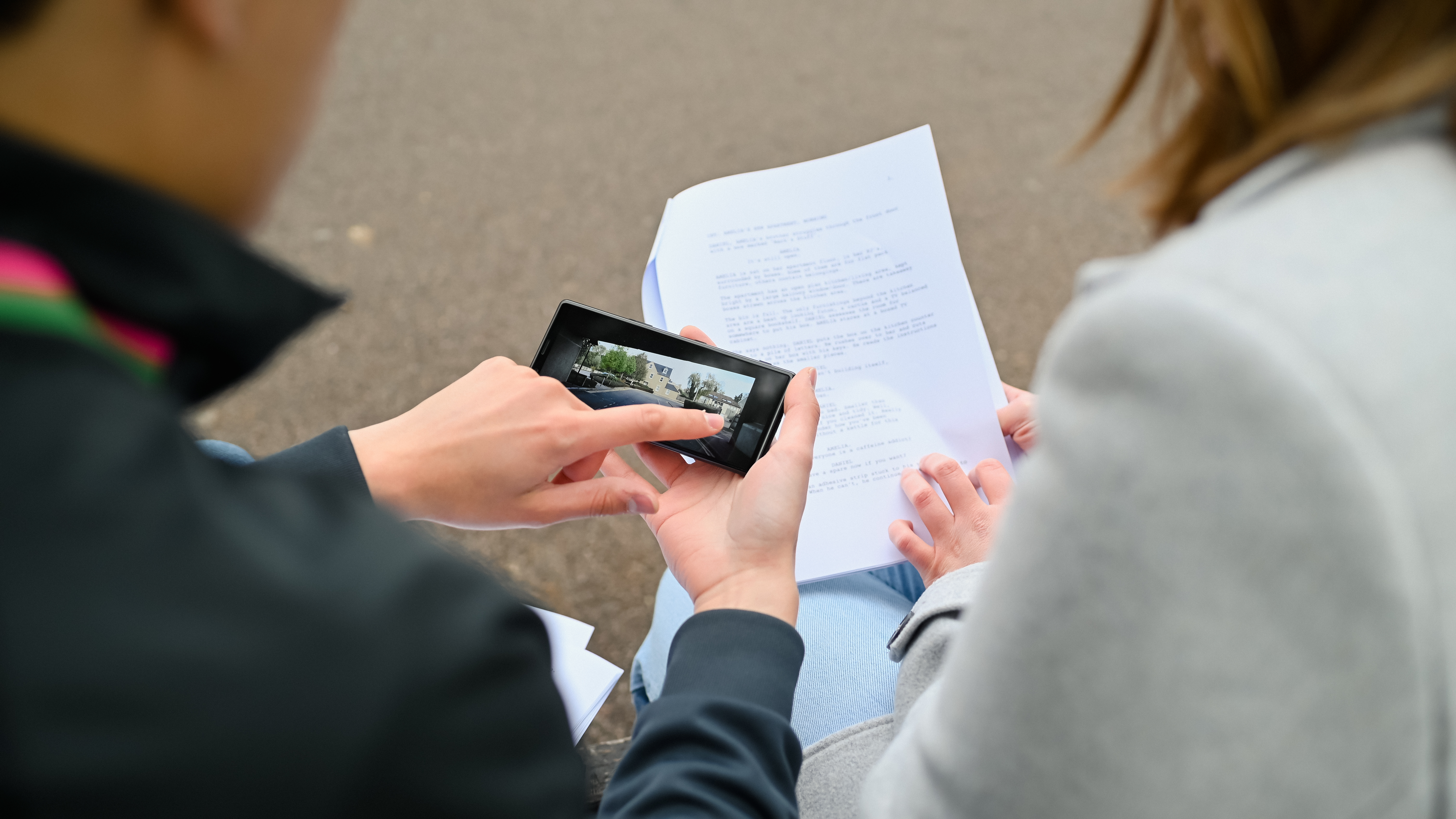 An over-the-shoulder view of a person holding a phone to review footage, while another consults a script.