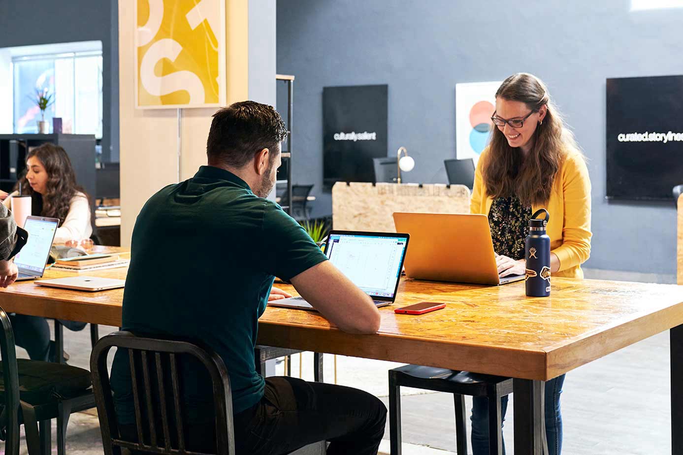 Três pessoas trabalham em laptops em uma mesa longa em um espaço de escritório comum
