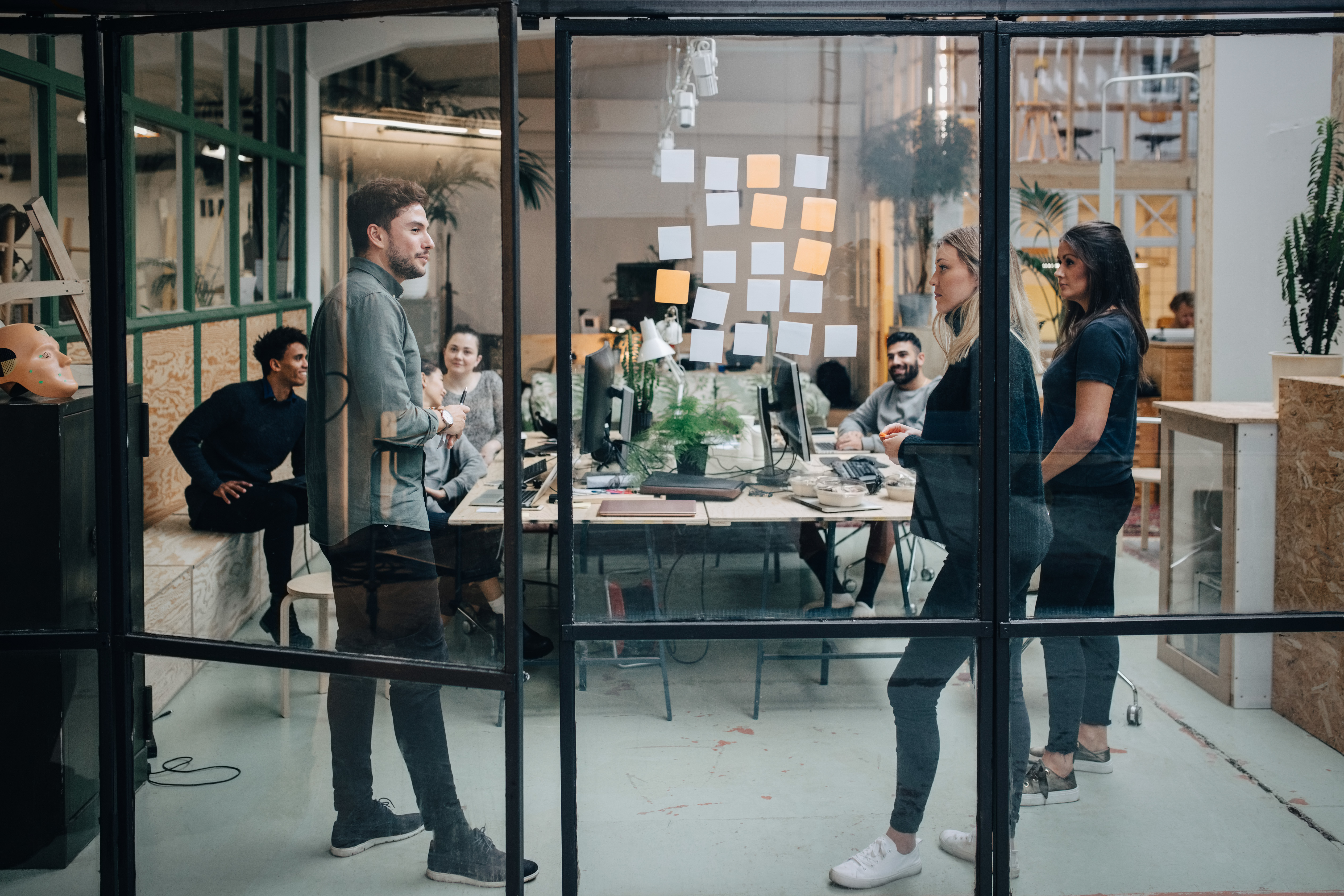 Colleagues look at a sequence of sticky notes on a glass wall in a modern office.