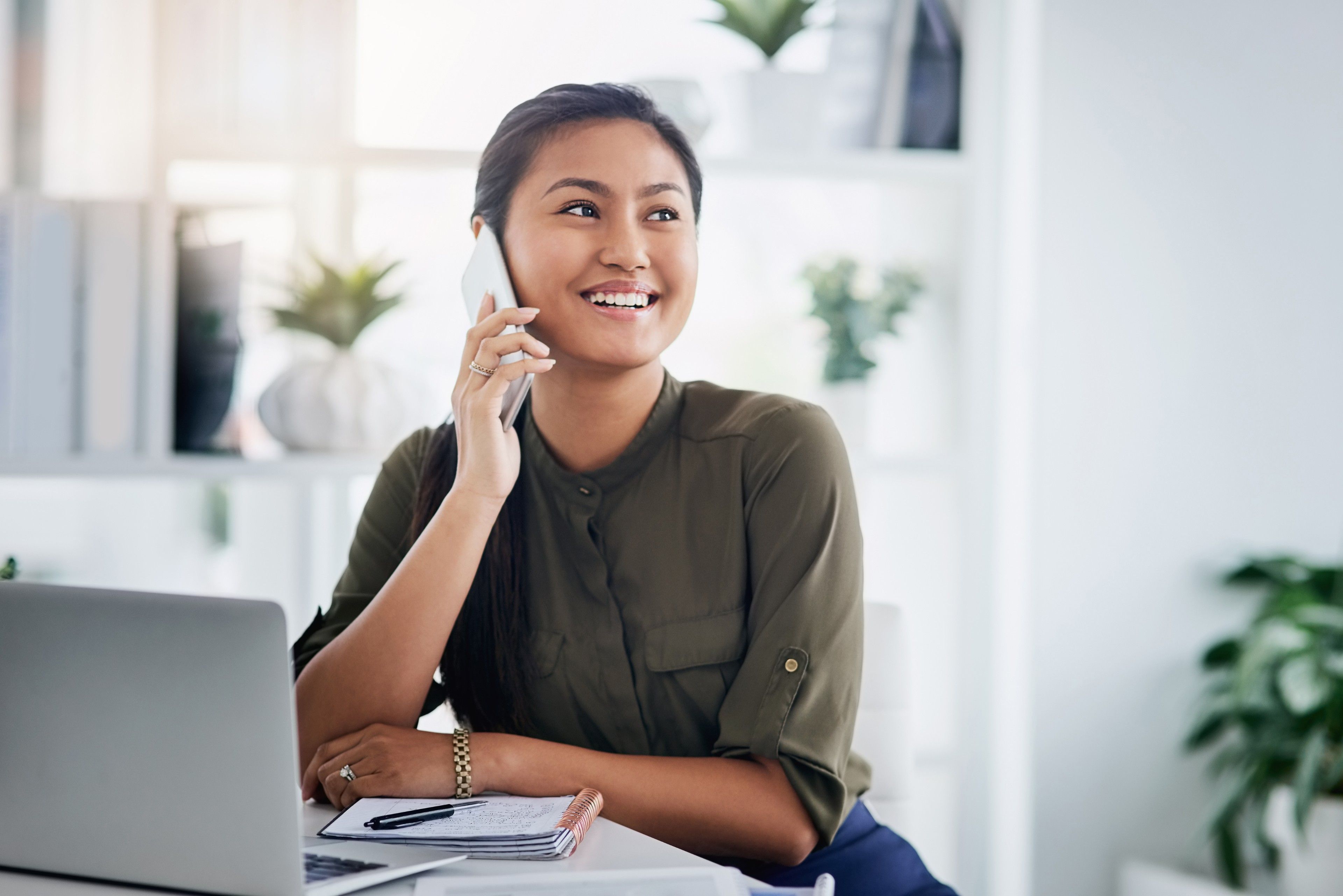 A person smiling as they sit at their laptop and enjoy a phone conversation.