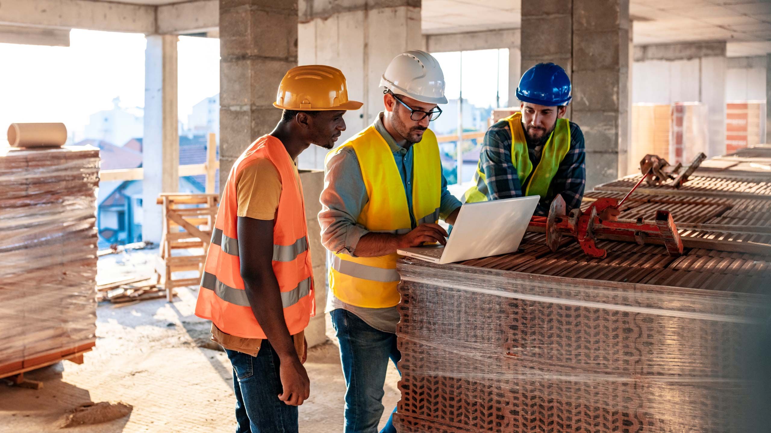 Três trabalhadores da construção civil usando capacetes e coletes de segurança olhando para um laptop