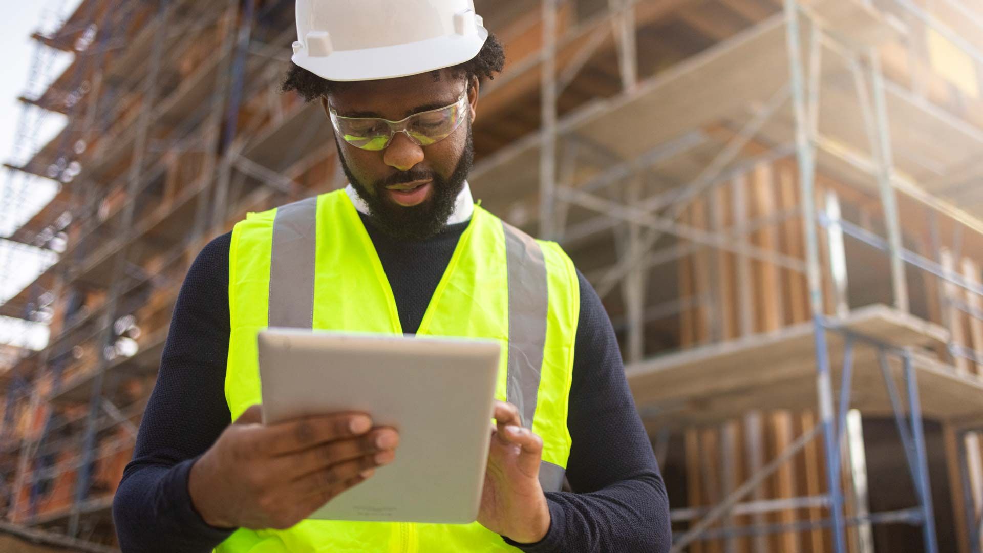 Un homme portant un casque de chantier, des lunettes et un gilet de sécurité, debout devant un bâtiment en construction