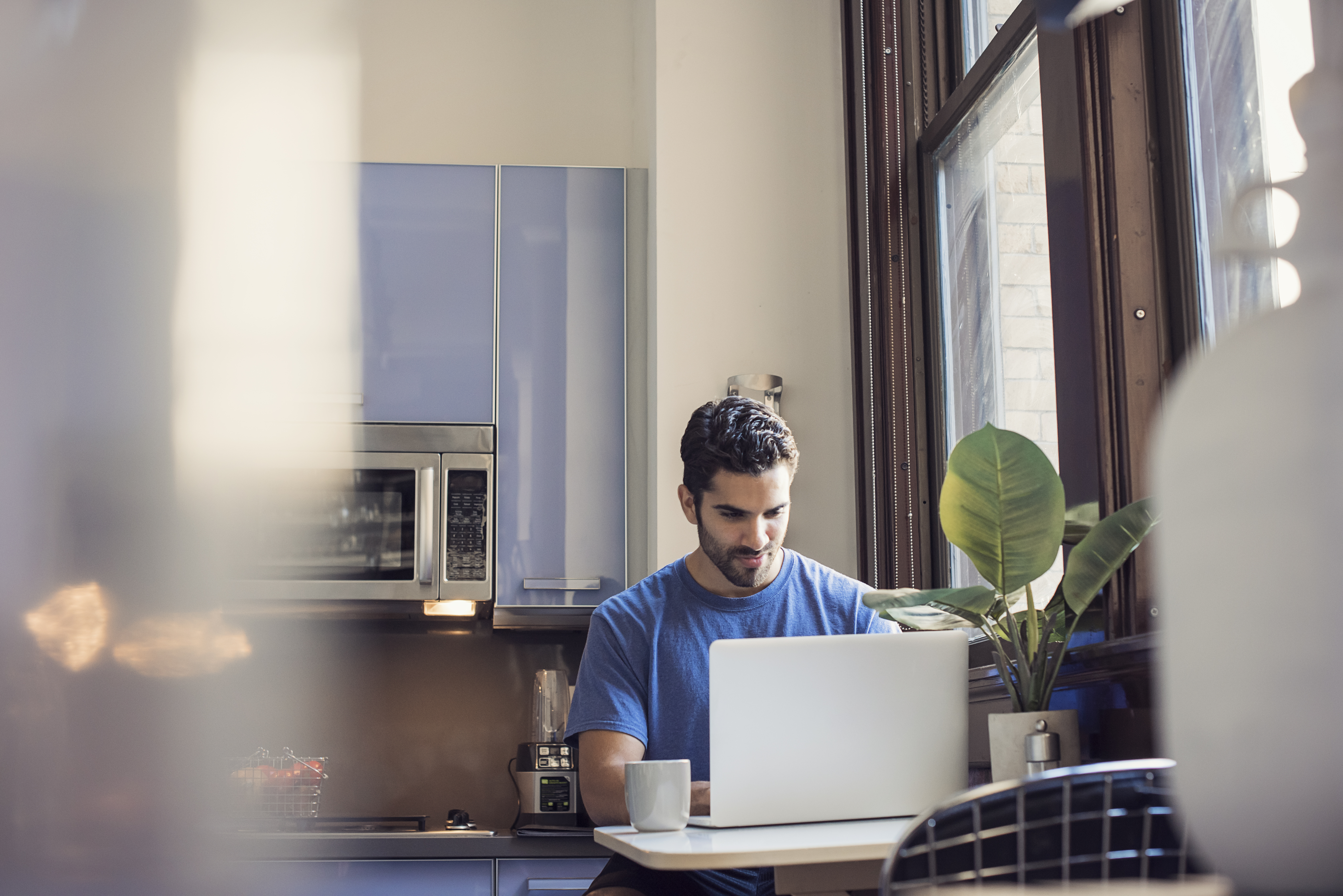 Person sitting at a table working on a laptop in a bright kitchen space with a cup of coffee nearby.