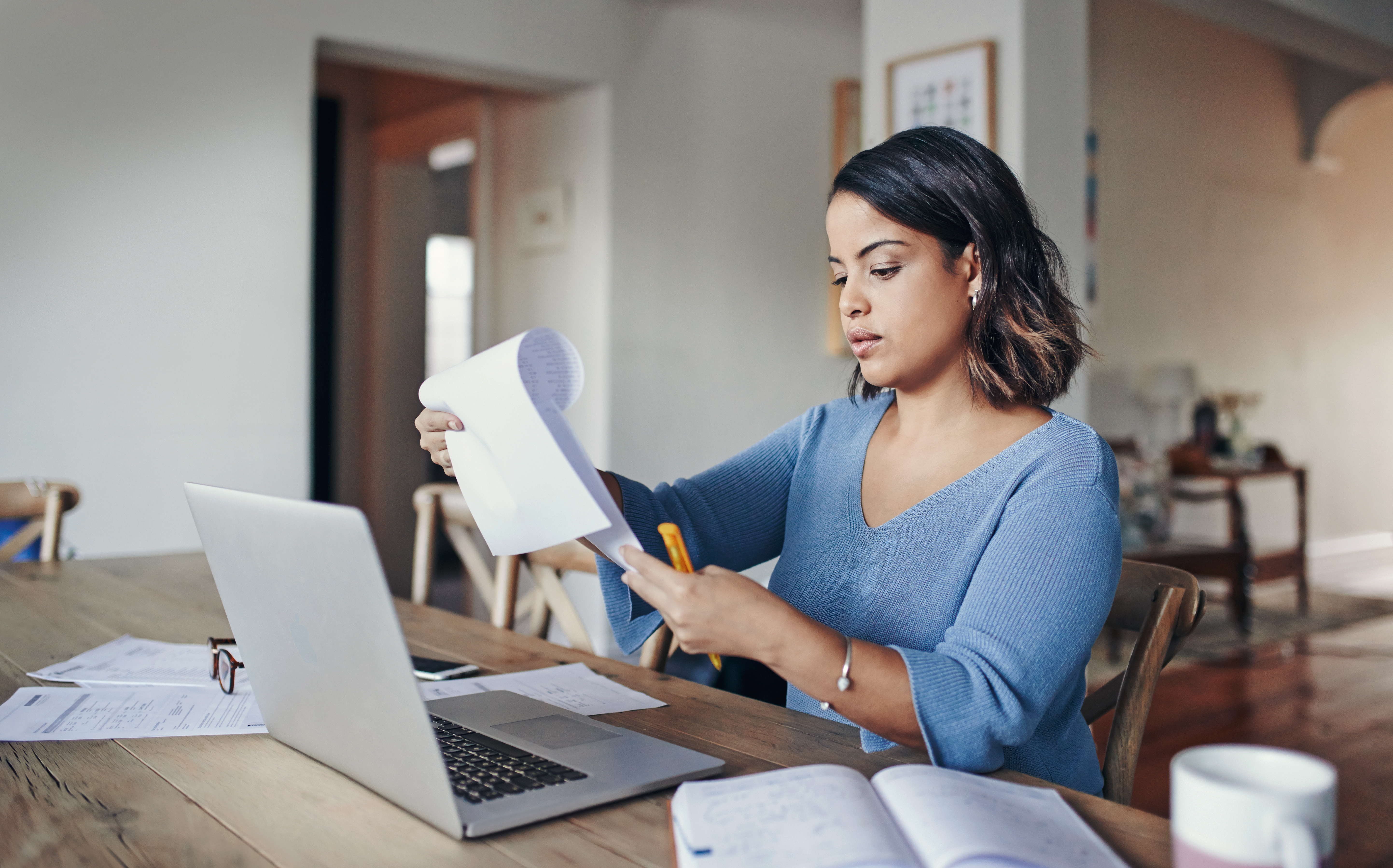 A remote worker looks at a paper document, while their laptop sits open on the table in front of them.