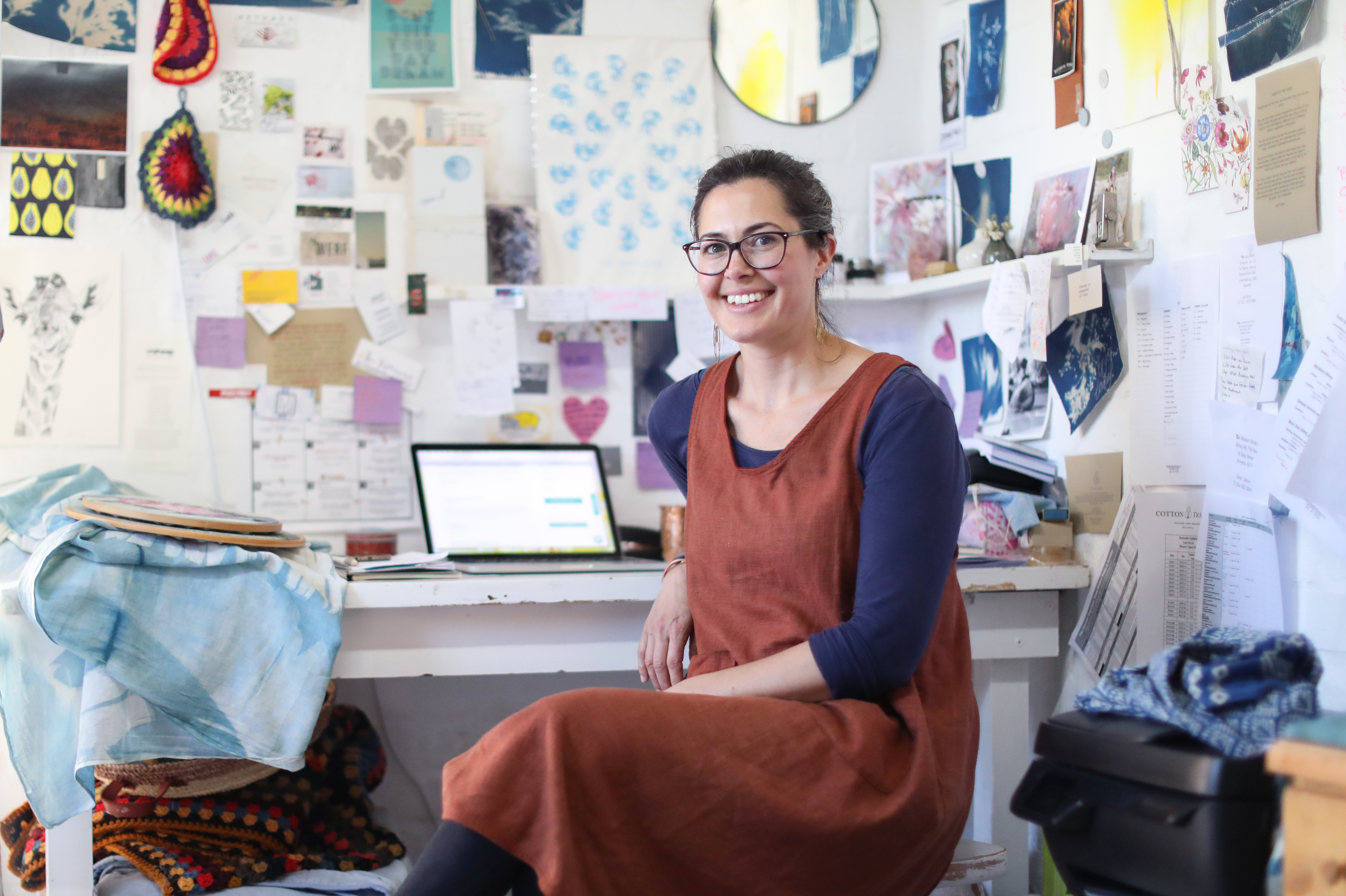 Person sitting at a cluttered desk in a creative workspace, with a laptop and various papers on the wall behind them.