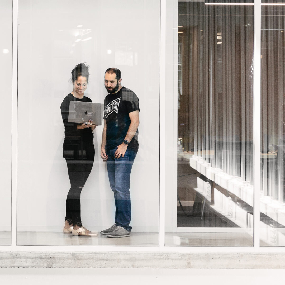 Dua orang berdiri memandang layar laptop di sebuah koridor gedung kantor